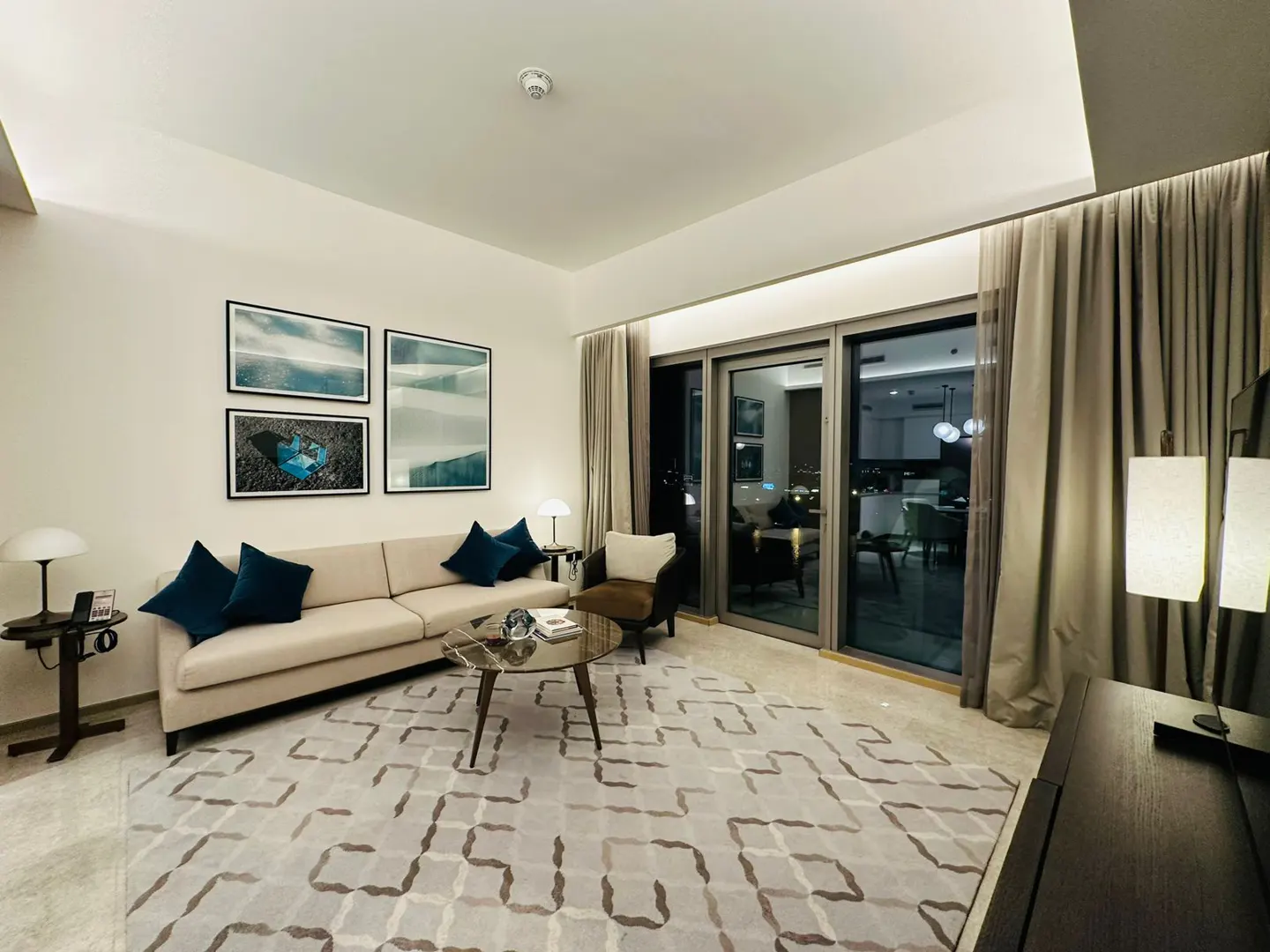 Living room with beige sofa, blue pillows, and abstract art. A patterned rug covers the floor, and sliding glass doors lead to a balcony.