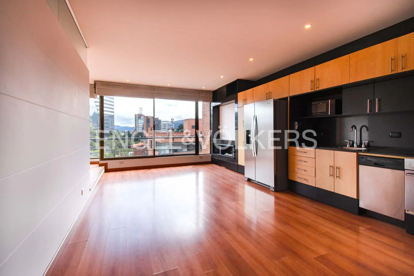 Bright, modern apartment with wood floors, a stainless steel fridge, and a large window overlooking a city.