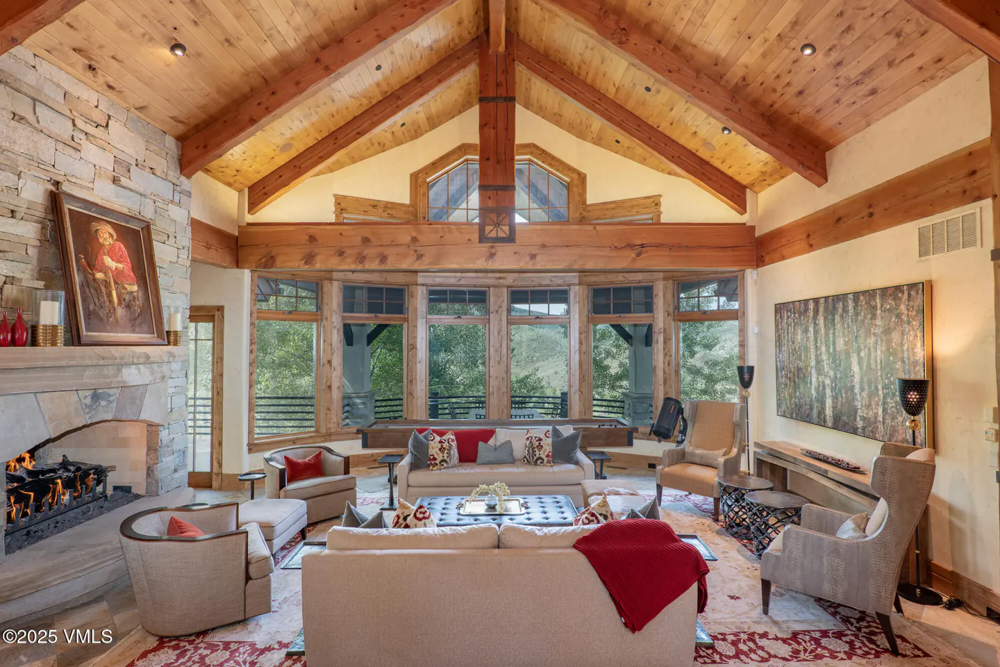 Living room with stone fireplace, wood ceiling beams, and large windows overlooking a green landscape. Neutral sofas and chairs.