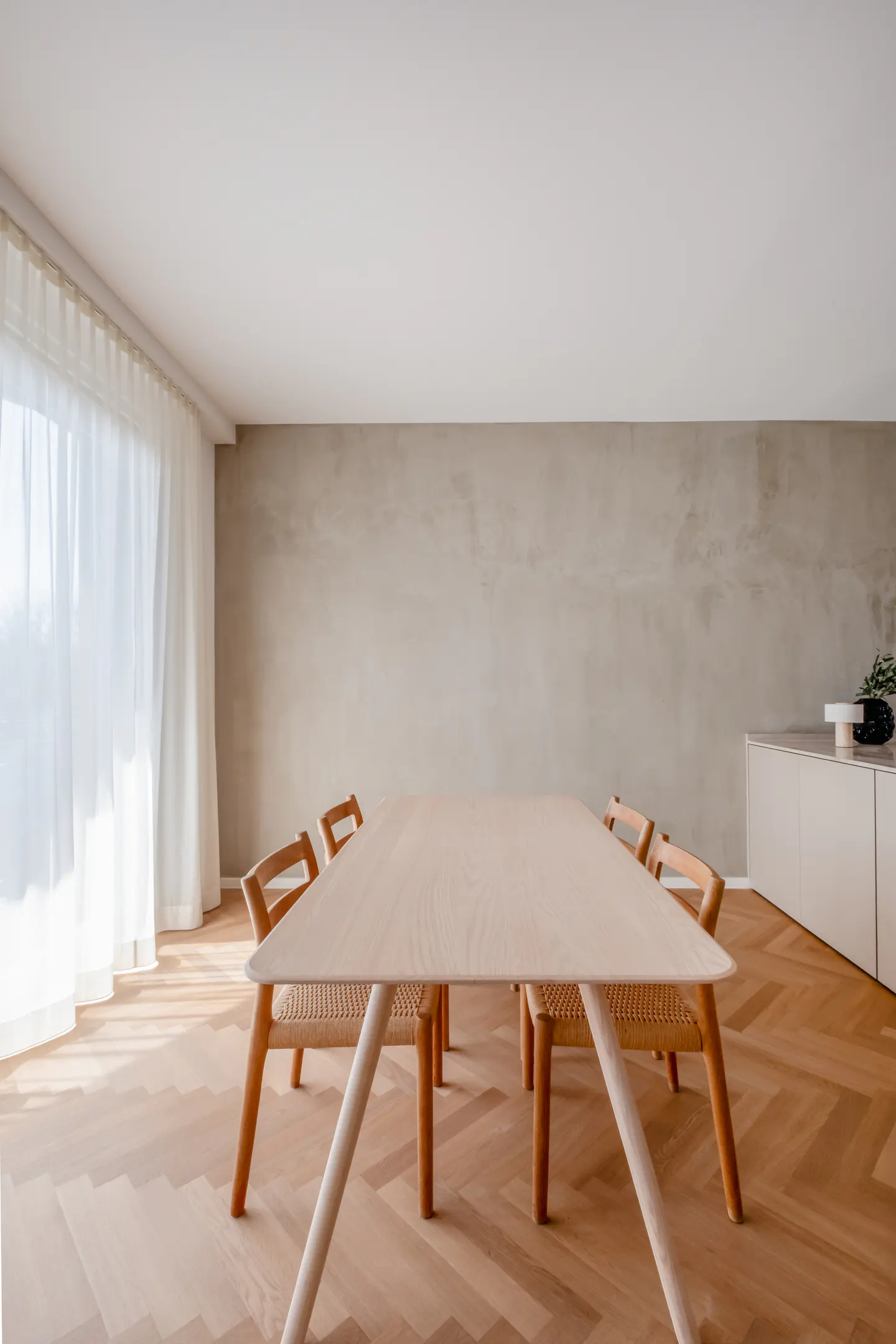Minimalist dining room with a light wood table, woven chairs, and herringbone wood floors. White curtains filter sunlight.