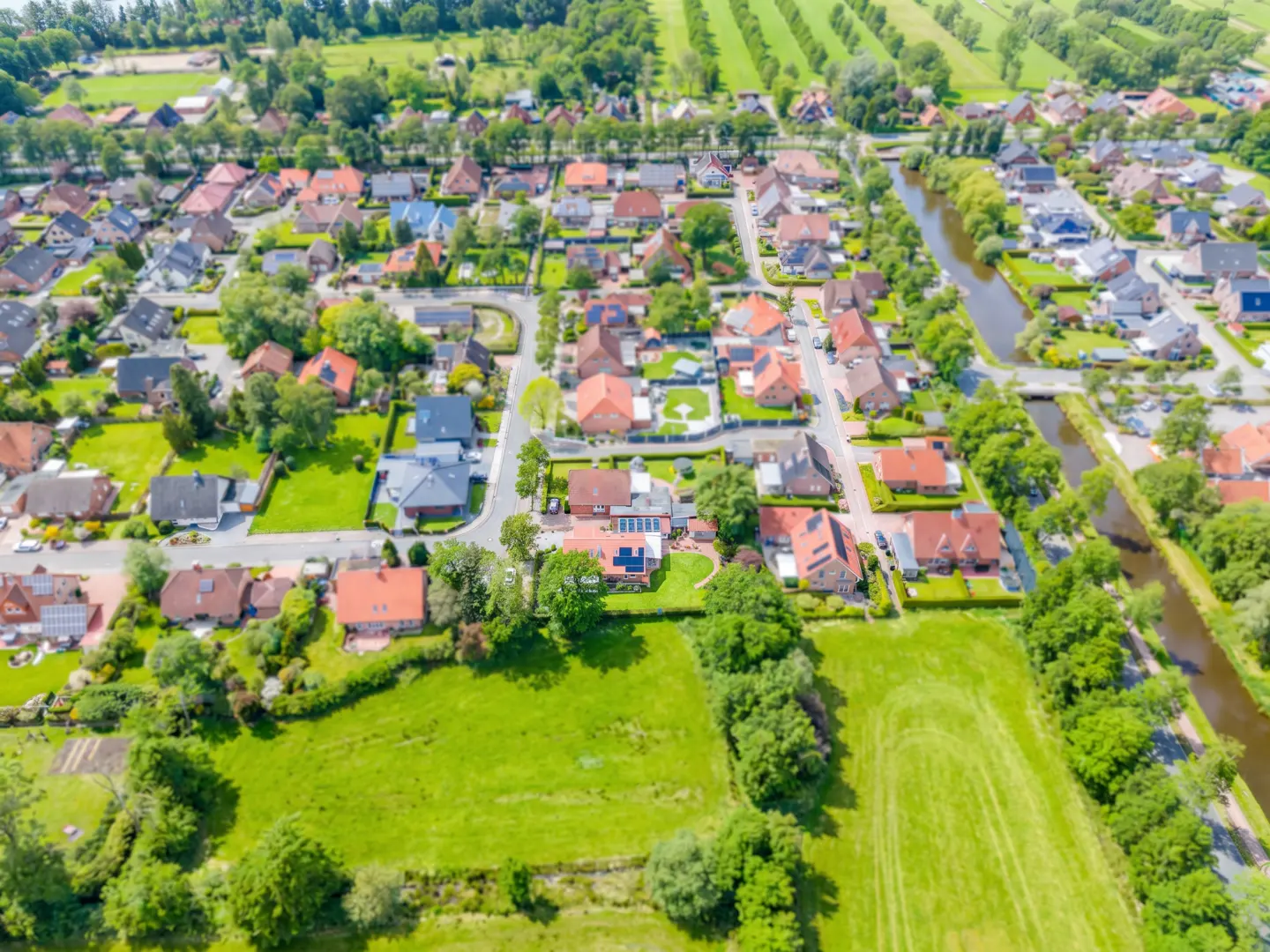 Aerial view of a quaint village with red-roofed houses, green lawns, and a winding canal under a sunny sky.
