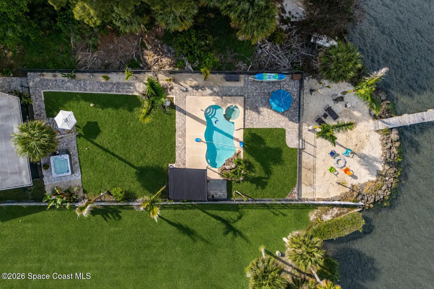 Aerial view of a waterfront property with a pool, lawn, and sandy beach area. Palm trees and lush greenery surround the property.