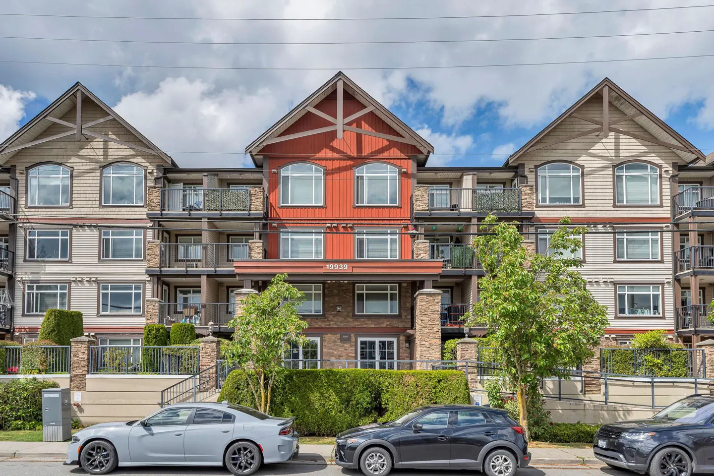 Exterior view of a three-story apartment building with balconies, cars parked in front, and green trees.