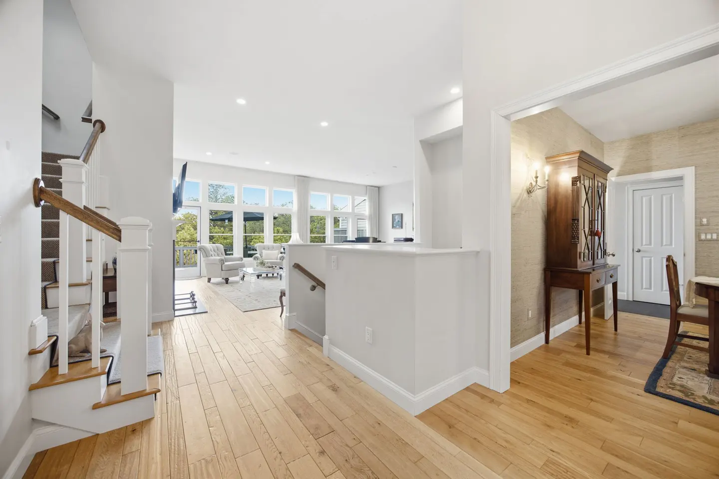 Bright home interior with hardwood floors, white walls, and a staircase. Living room visible with large windows and furniture. Dining room with hutch seen through doorway.