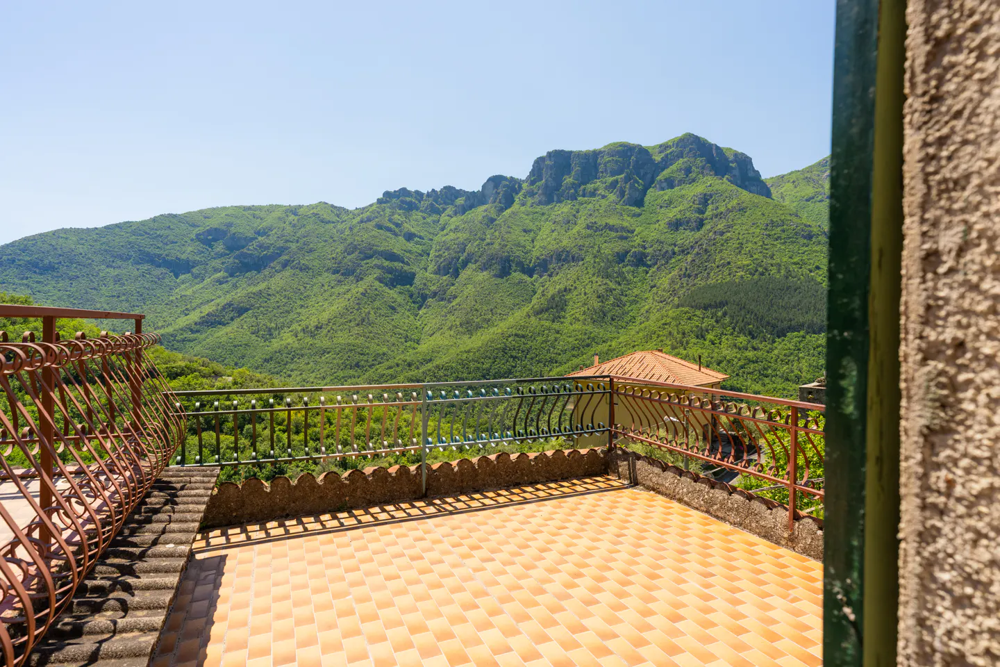 Tiled balcony with ornate metal railings overlooking a lush green mountain range under a clear blue sky.