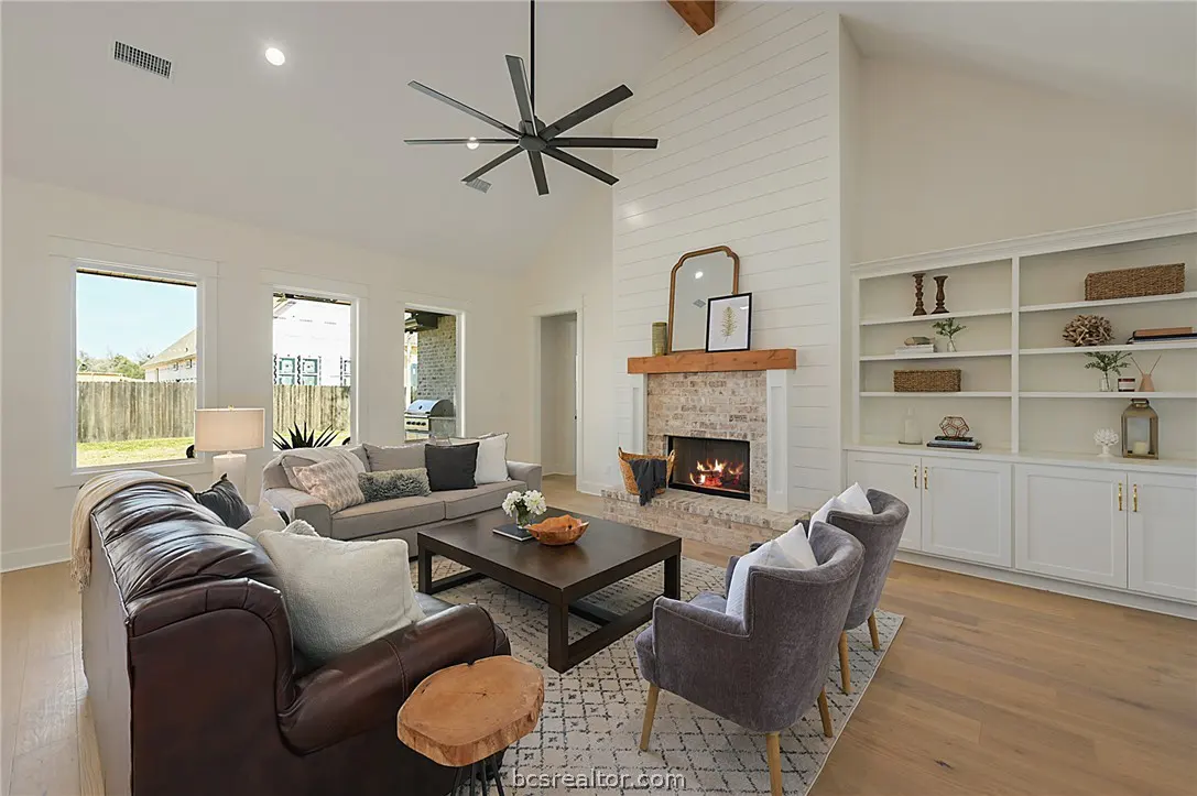 Bright living room with a brick fireplace, white shiplap wall, dark leather sofa, gray armchairs, and built-in shelving.