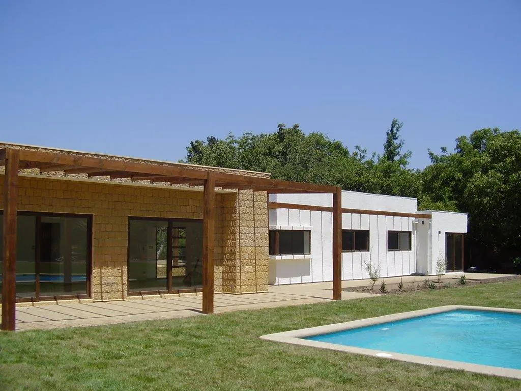 Modern home exterior with a pool, stone and white walls, and a wooden pergola under a clear blue sky.