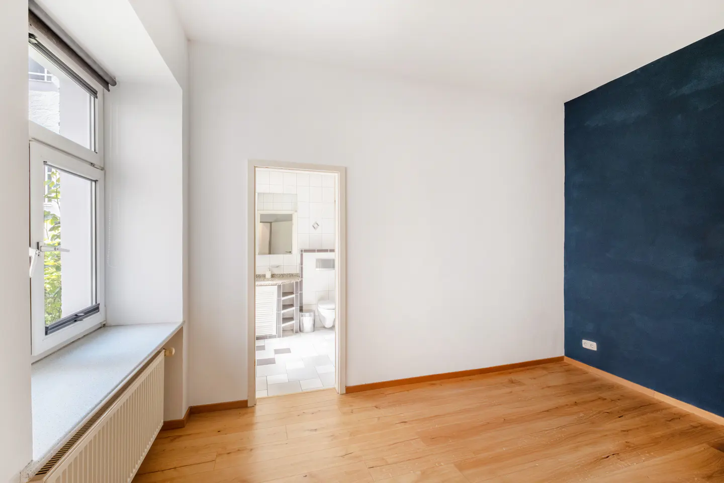 Bright room with wood floors, white walls, and a blue accent wall. A window with a radiator sits to the left, and a bathroom is visible through a doorway.