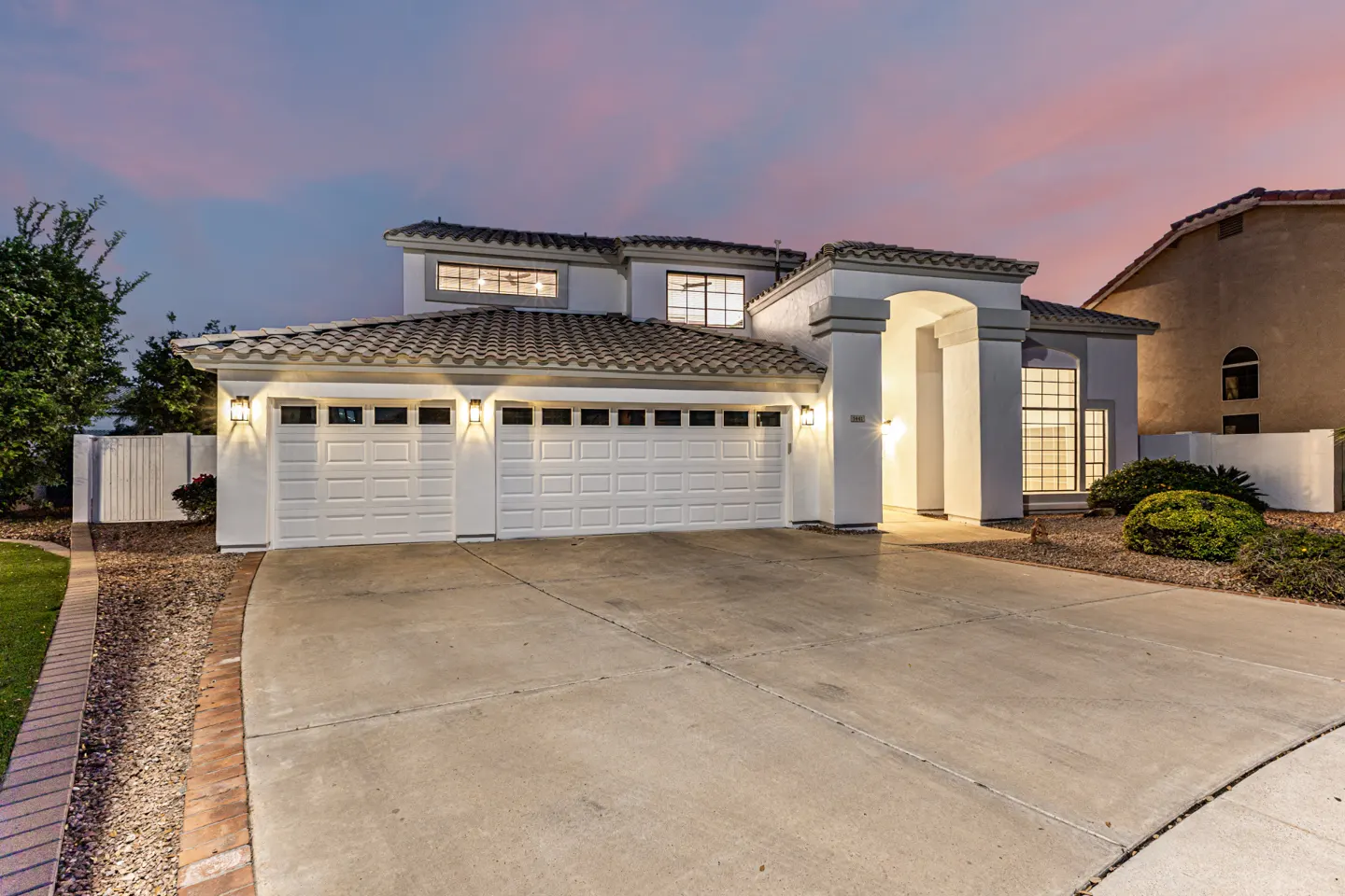 Two-story white house with a three-car garage, tile roof, and concrete driveway at dusk.