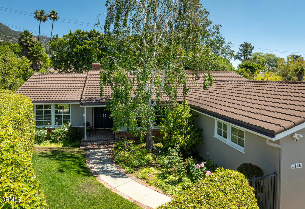 Front exterior of a single-story home with a brown tile roof, gray siding, and lush green landscaping. A birch tree stands in the front yard.
