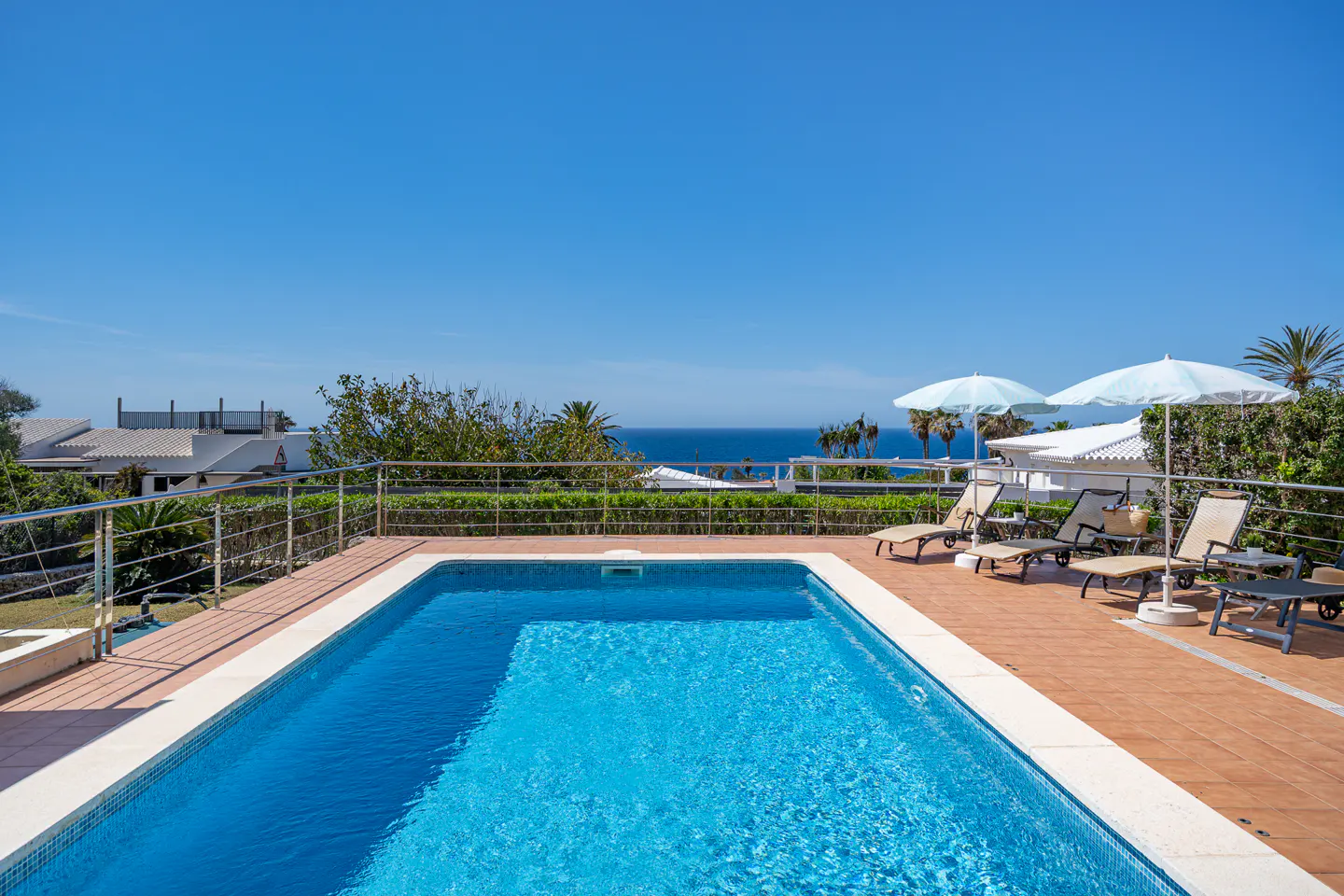 A rectangular blue swimming pool with lounge chairs and umbrellas on a brick patio overlooking the ocean.