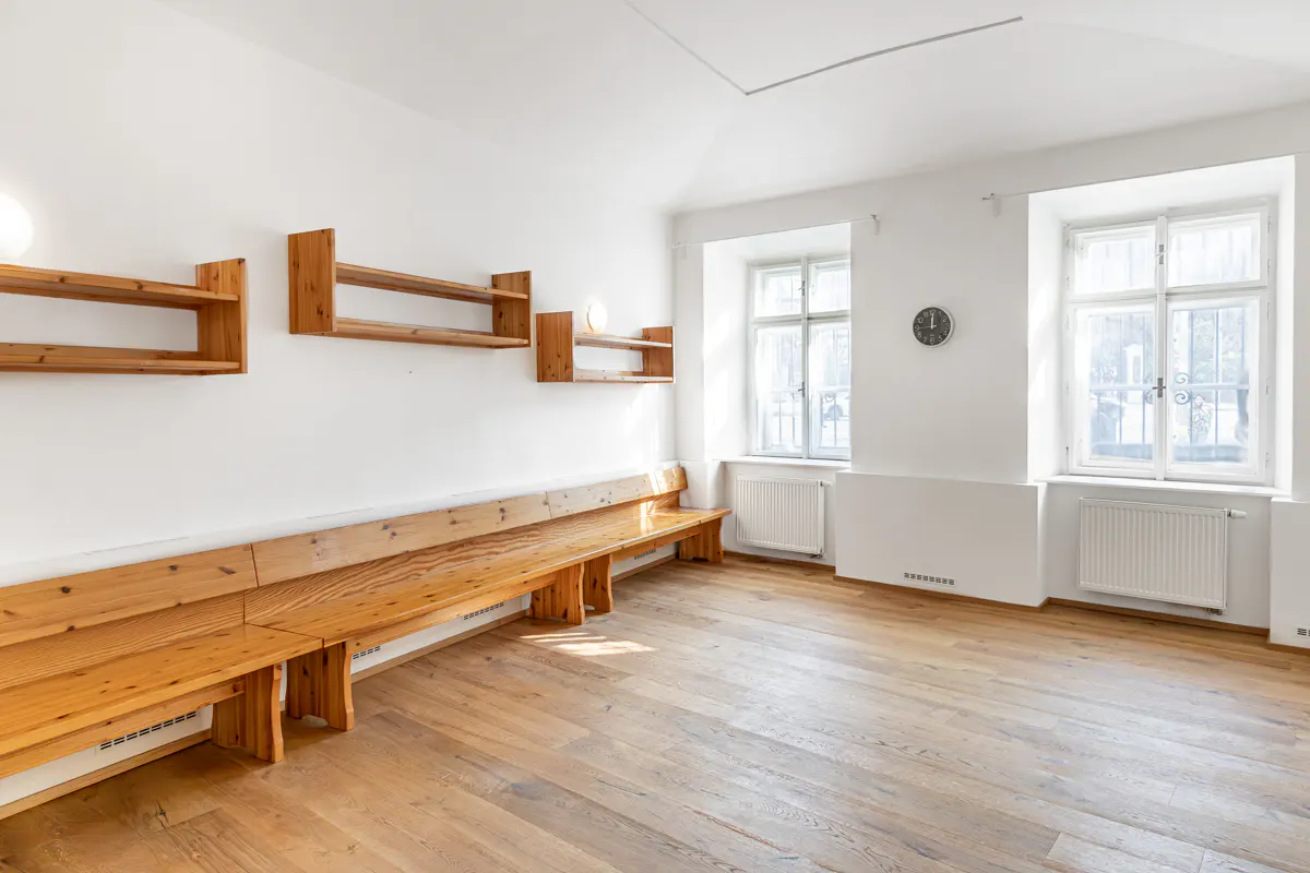 Bright, empty room with wood floors, white walls, and natural light. Wooden benches line the wall under floating shelves. Two windows are visible.