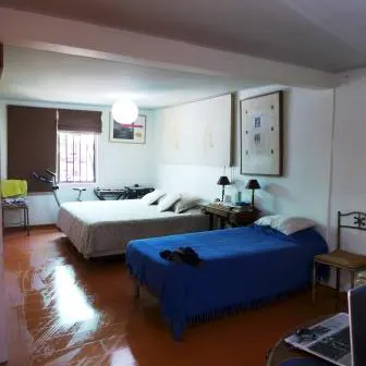 Bedroom with two beds, one white and one blue, on a shiny orange floor. There is a window with brown blinds and exercise equipment.