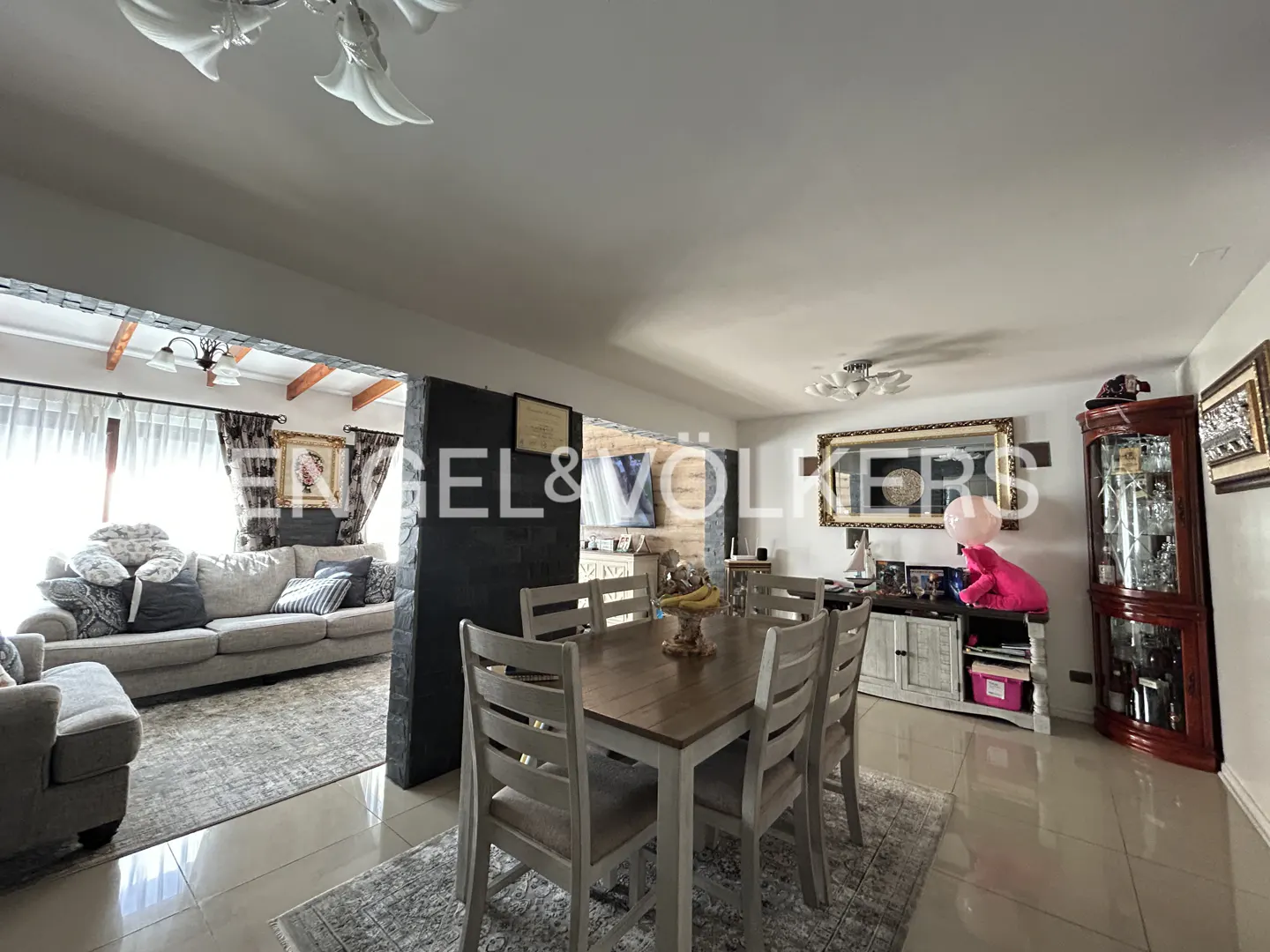 Open-concept living and dining area with a wooden table, gray chairs, and a beige sofa. Tiled floors and neutral color palette.