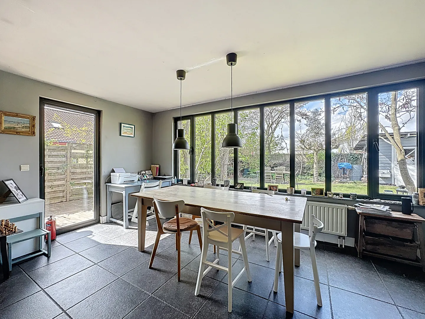 A dining room with gray walls, black tile floors, and a large window overlooking a green backyard. A long wooden table with chairs sits under pendant lights.
