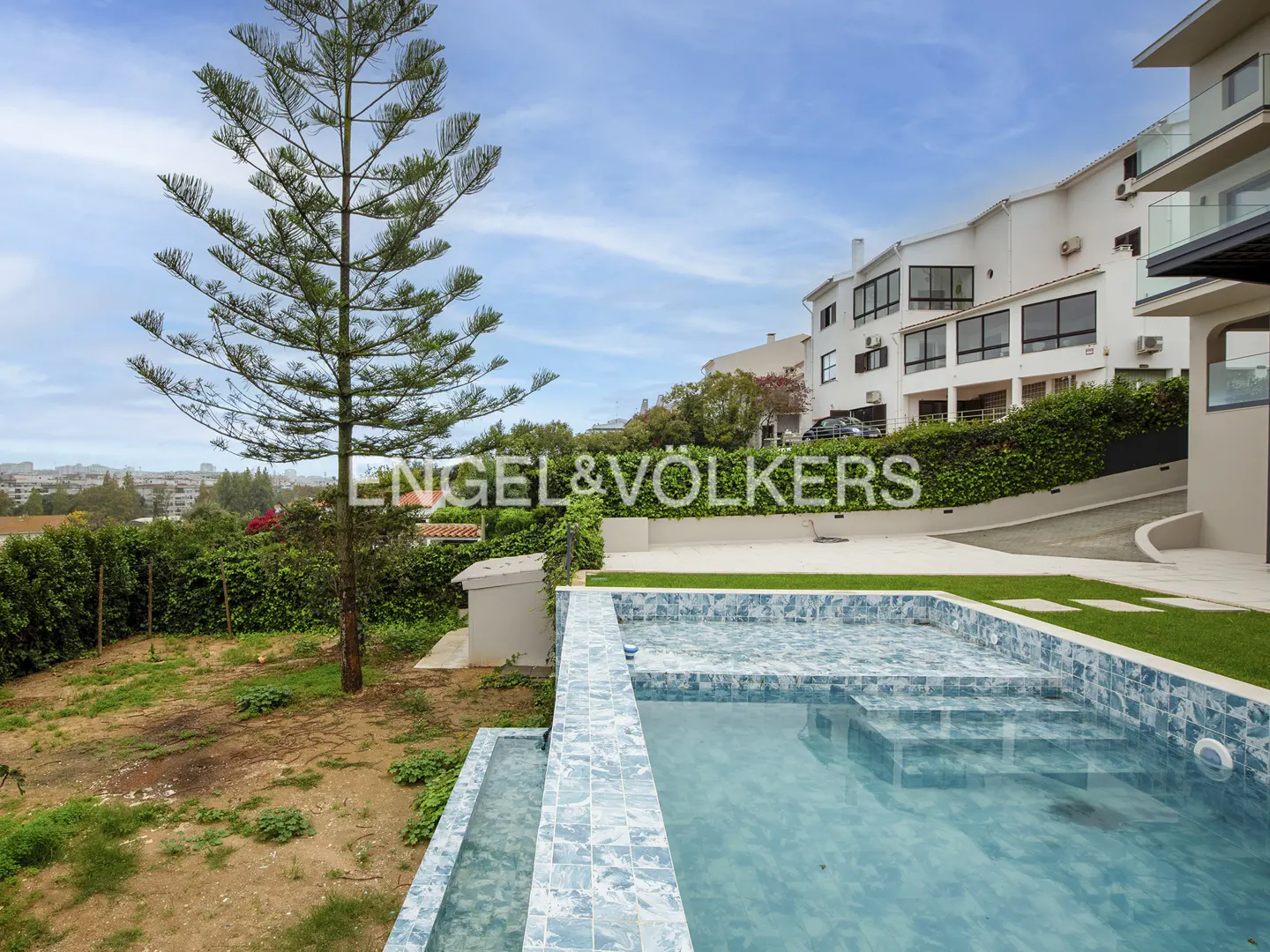 Backyard view of a pool with blue tiles, a tree, and a white building in the background.