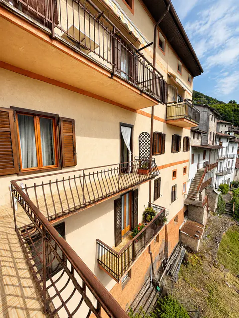 Exterior view of a multi-story building with balconies and brown shutters, overlooking a hillside village under a blue sky.