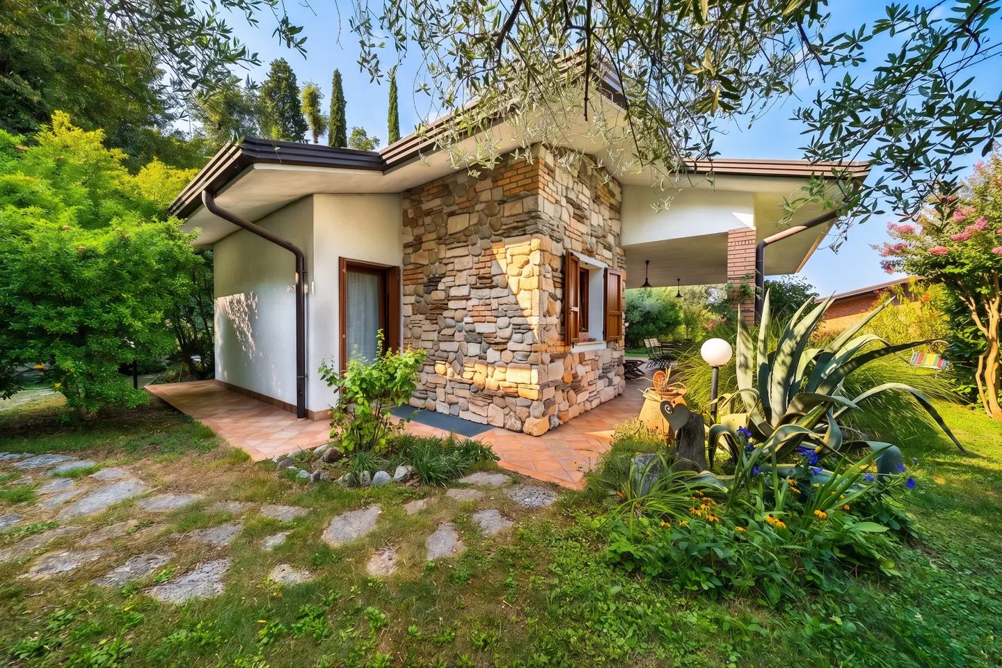 Exterior view of a stone and stucco house with a brown roof, surrounded by green grass and plants.