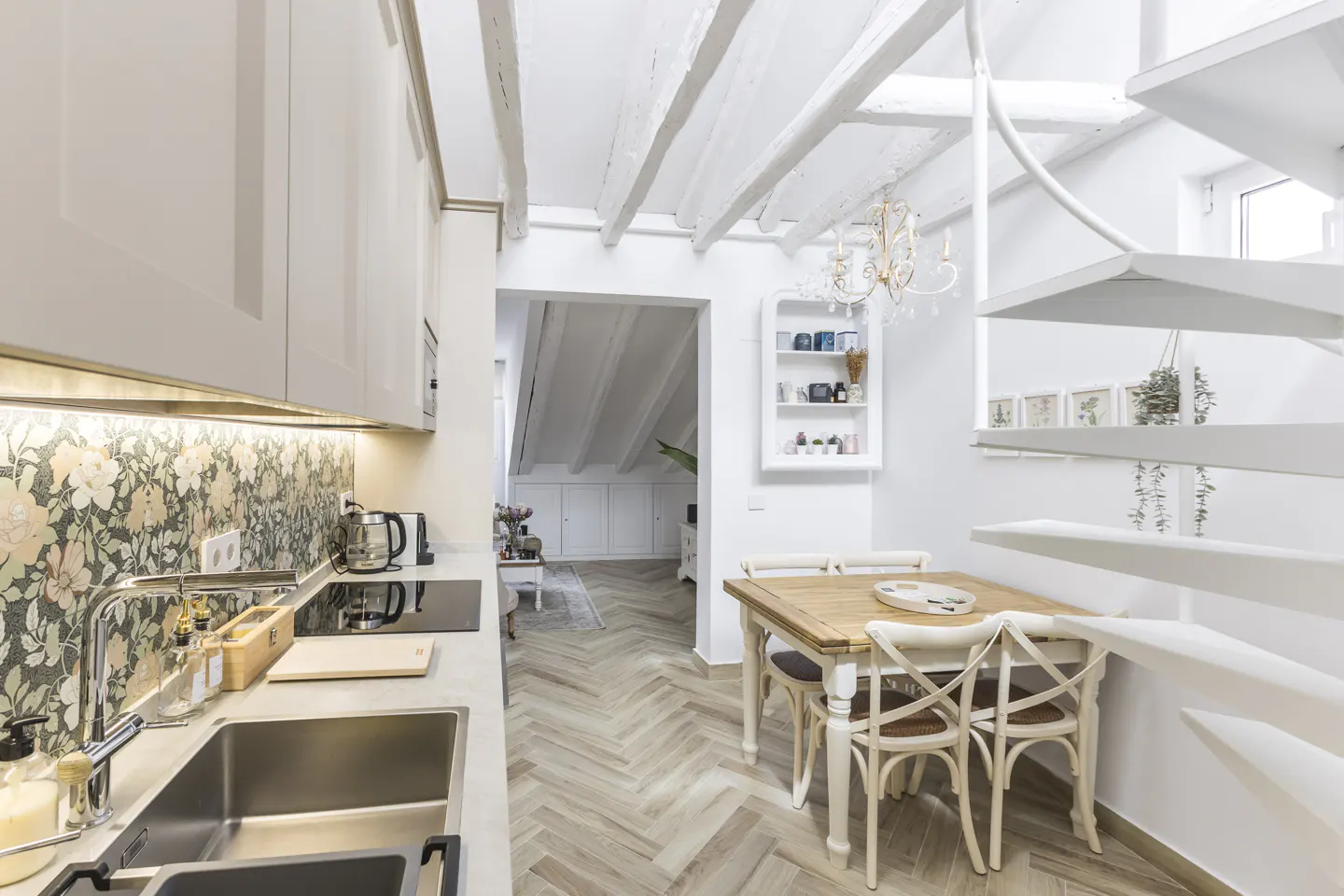 Bright, modern kitchen with white cabinets, floral backsplash, and stainless steel sink. A wooden table with chairs sits near a white spiral staircase.