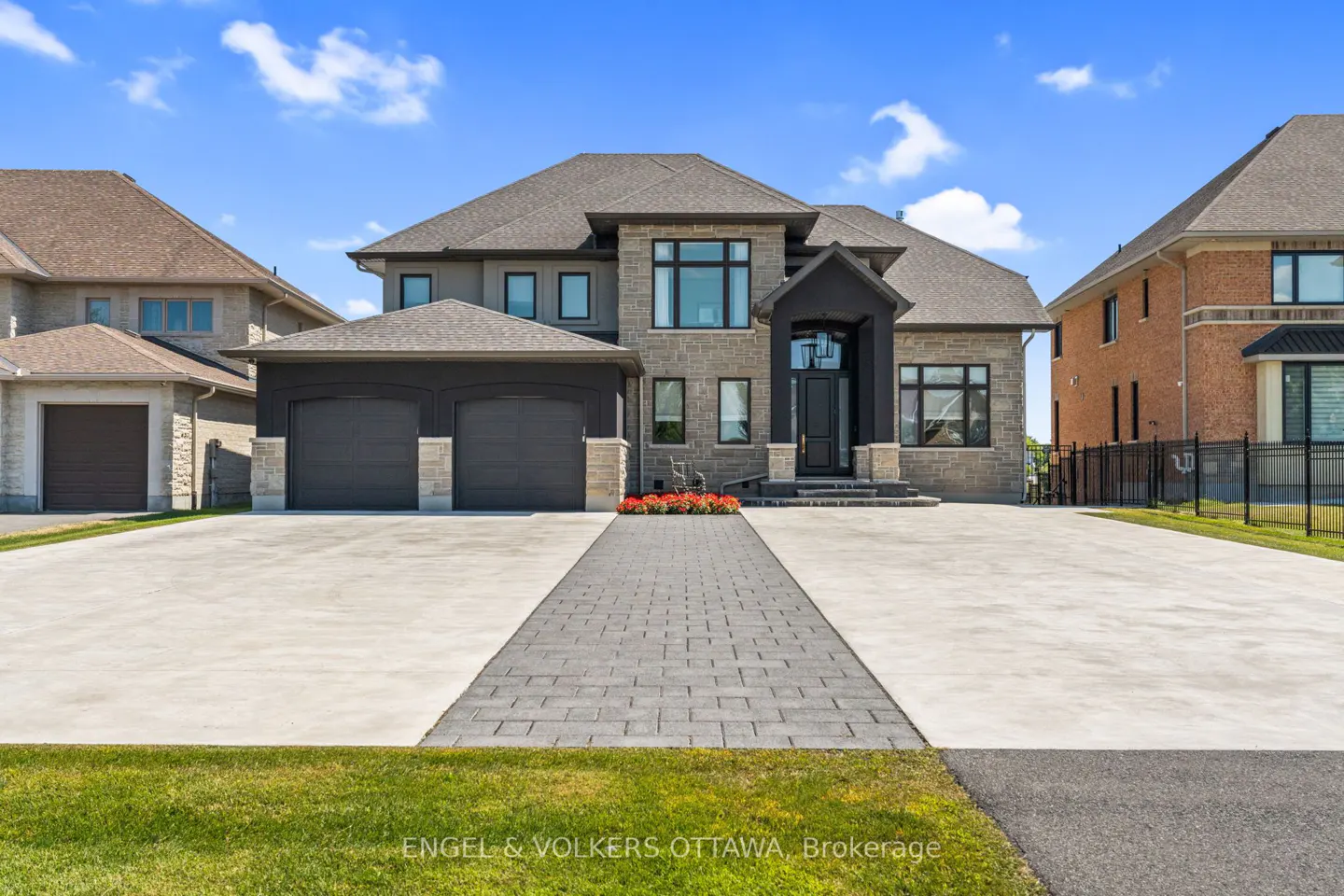 Two-story stone house with a gray roof, black trim, and a long driveway on a sunny day.