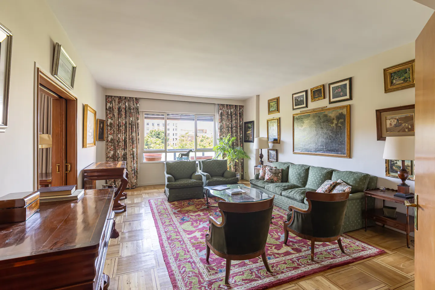 Living room with green sofa and chairs, a red patterned rug, and many framed pictures on the wall. A large window shows a city view.