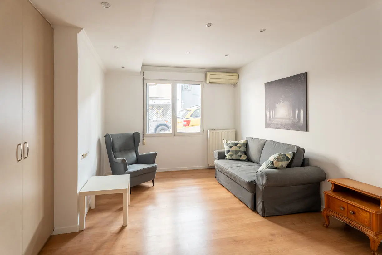 Bright living room with wood floors, gray sofa and armchair, white walls, and a window. A wooden cabinet sits on the right.