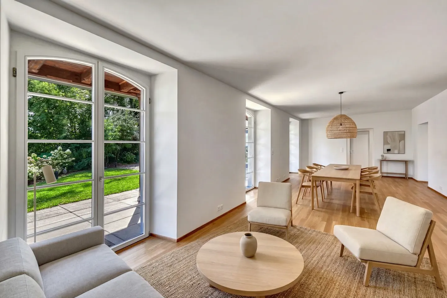 Bright, open-concept living and dining area with hardwood floors and white walls. Large windows overlook a green lawn. Neutral-toned furniture.