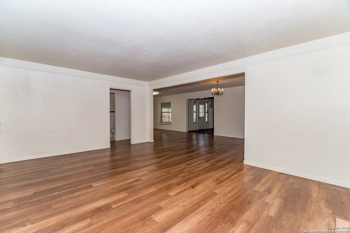 Spacious living area with white walls, wood-look flooring, and an open floor plan leading to a dining room with a chandelier.