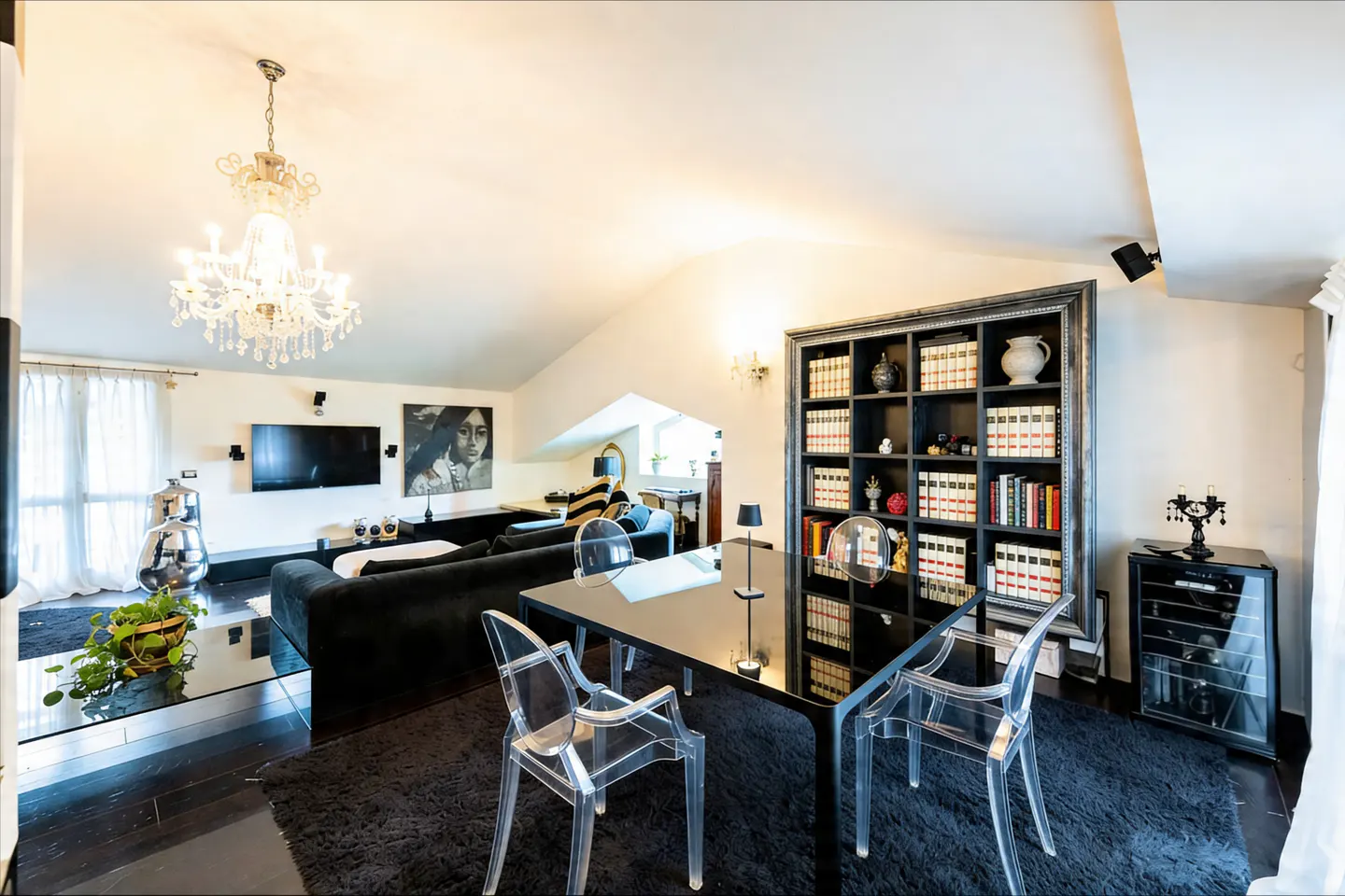 Living room with black furniture, a crystal chandelier, and a black bookcase filled with books. Clear acrylic chairs surround a black table.