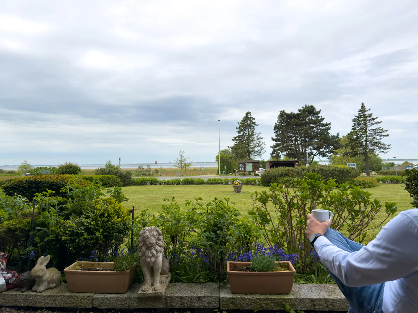 A person sits with a coffee cup overlooking a green lawn, trees, and a distant body of water under a cloudy sky. Statues and planters line the foreground.