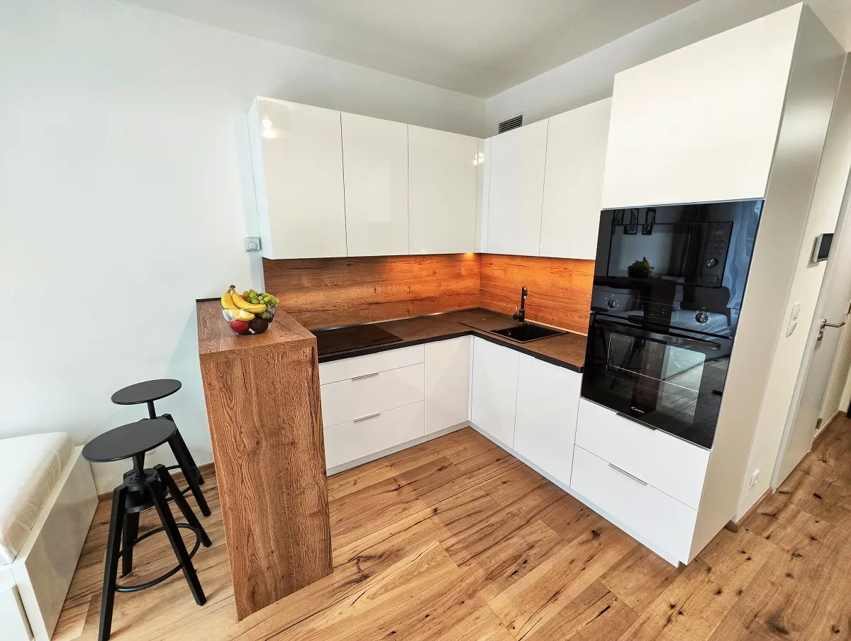 Modern kitchen with white cabinets, wood countertop and backsplash, black sink and oven, and wood floors. Two black stools sit by a wood counter.