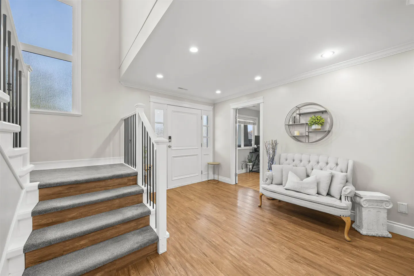 Bright foyer with wood floors, white walls, and stairs. A gray tufted settee sits near a round shelf. A white door is centered.