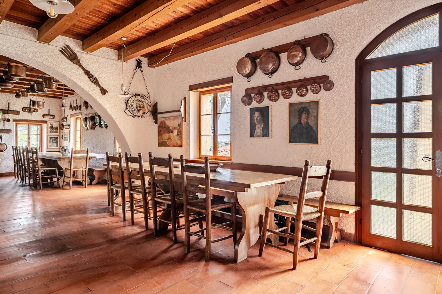 Rustic dining room with long wooden tables, chairs, and terracotta tile floor. Copper pots hang on the white textured walls. A large fork decorates the archway.