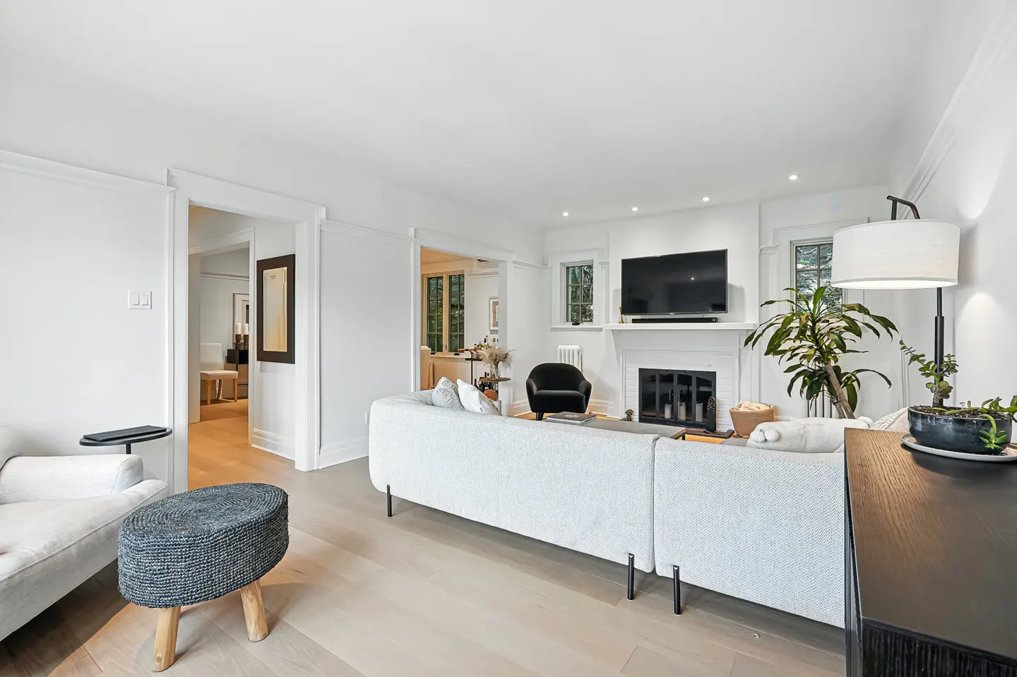 Bright living room with white walls, light wood floors, and a large gray sectional sofa facing a fireplace with a TV above it.