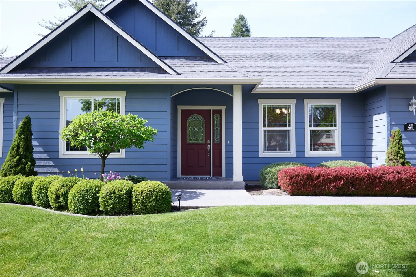 Blue house with a red door, white trim, and green lawn. Bushes and a small tree are in front of the house.