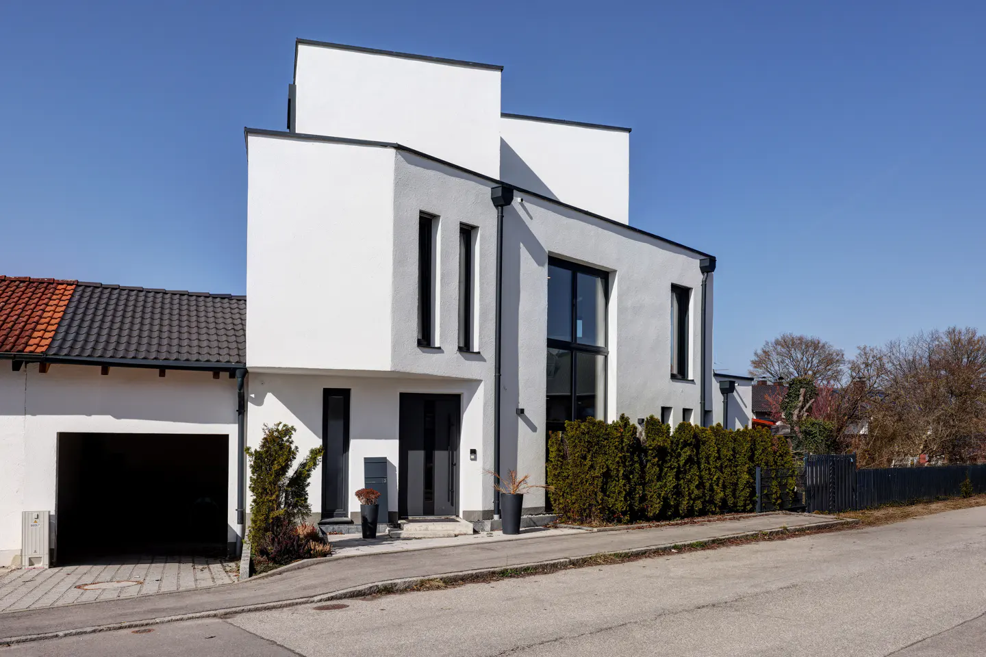 Modern two-story house with white and gray exterior, black trim, and a hedge-lined front yard on a sunny day.