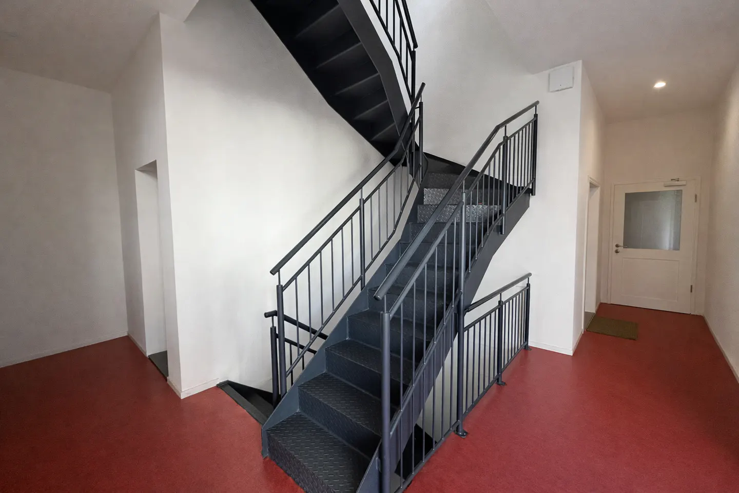Interior view of a building with a black metal staircase, white walls, and a red floor. A white door is visible in the background.