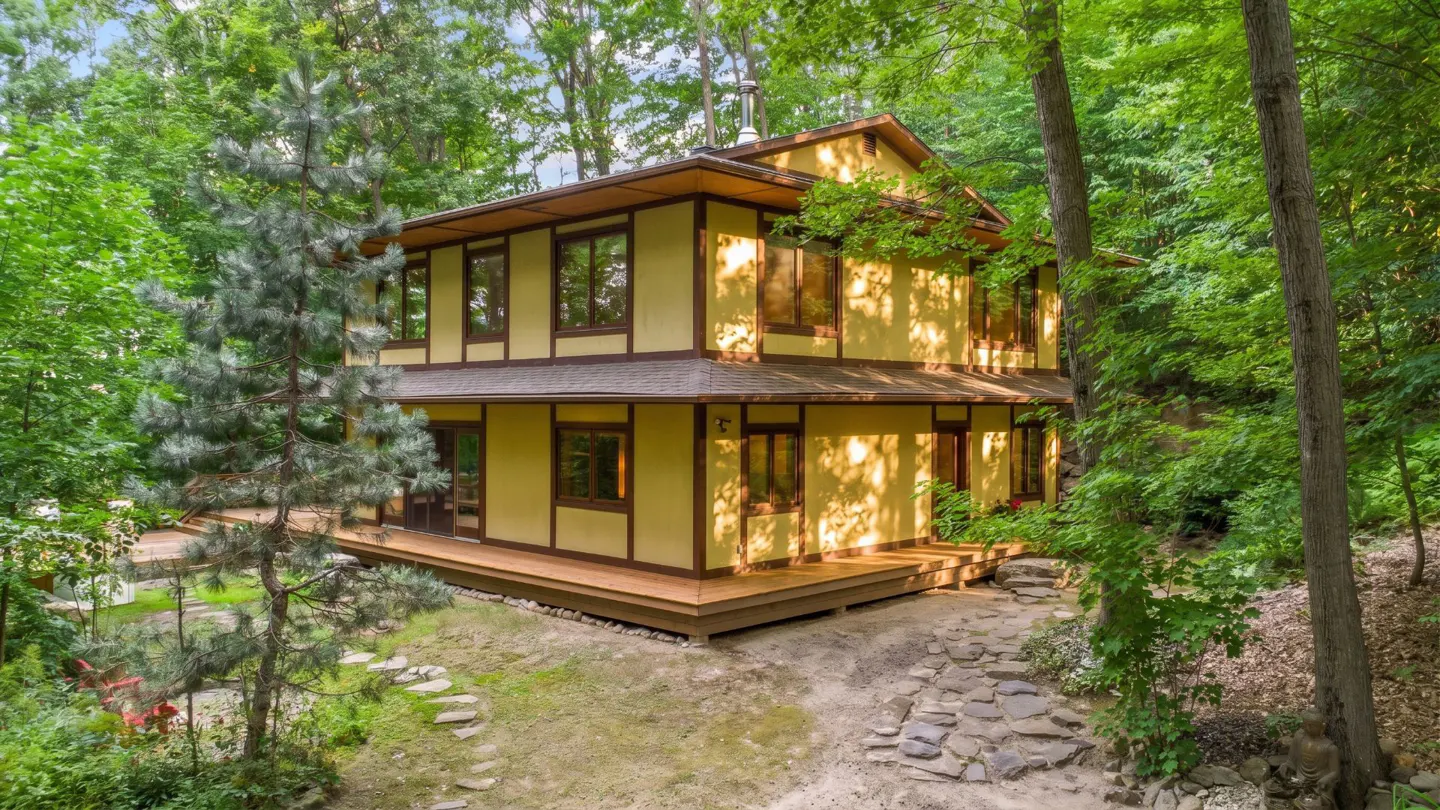 Two-story yellow house with brown trim and a wooden deck, surrounded by lush green trees. A stone path leads to the house.