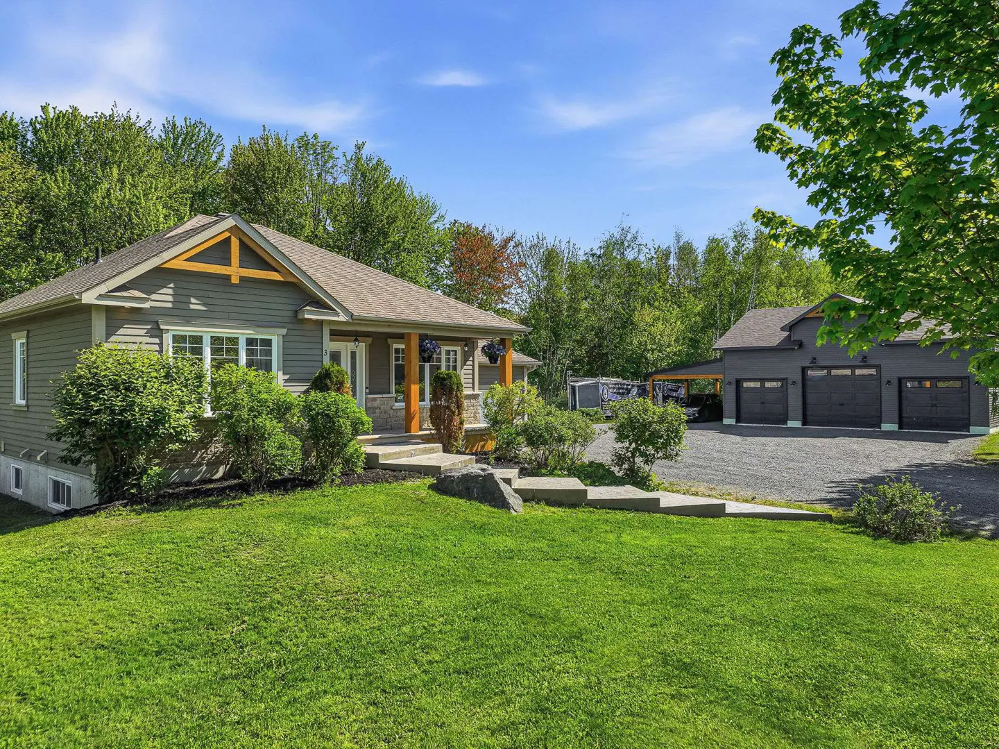 A grey bungalow with wood accents, a green lawn, and a detached three-car garage on a gravel driveway under a blue sky.