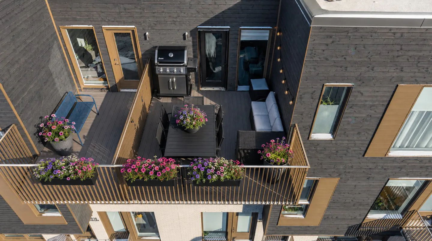 Aerial view of a modern rooftop deck with a grill, dining table, sofa, and colorful flower boxes. The building has dark wood siding and light wood trim.