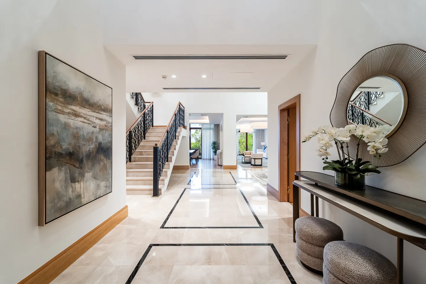 Bright foyer with marble floors, abstract art, and a staircase. A console table with orchids and a decorative mirror are on the right.