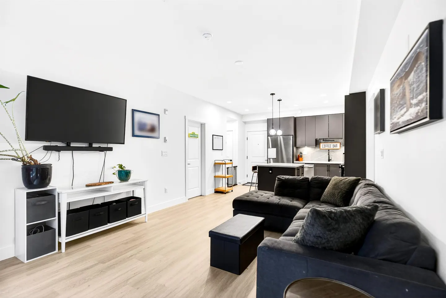 Bright, open-concept living room with a large black sectional sofa, TV, and modern kitchen in the background.