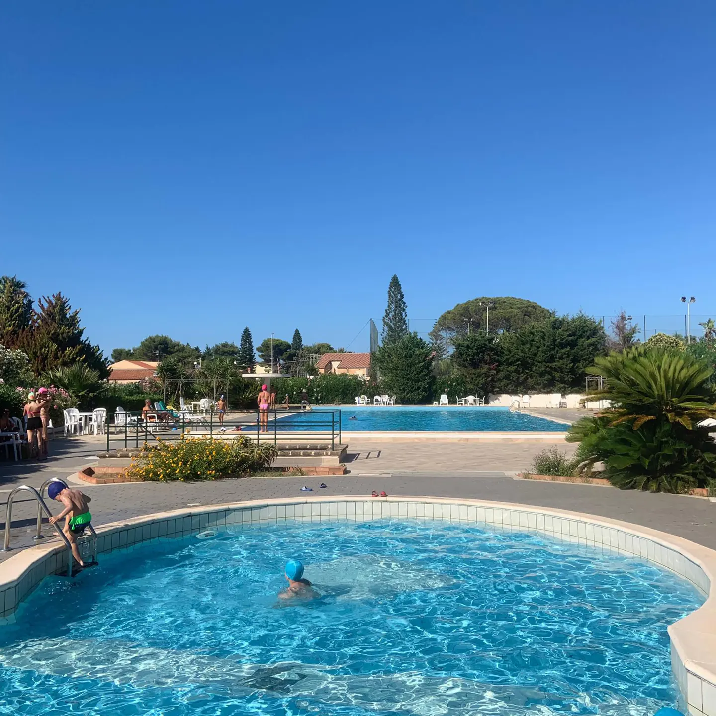 Outdoor pool area with people swimming and lounging under a clear blue sky.