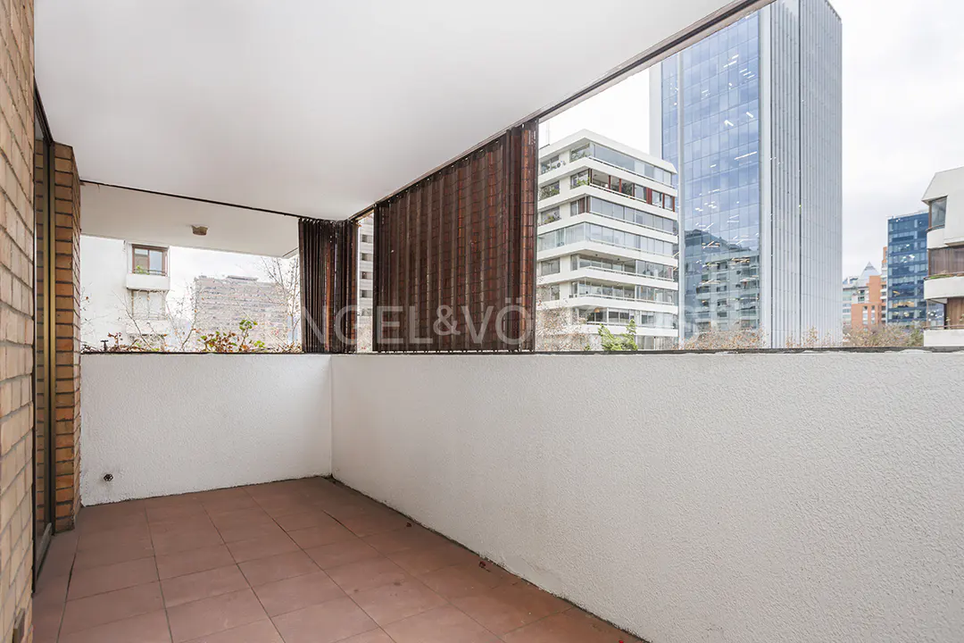 A balcony with brown tile flooring and a white wall overlooks city buildings. A brown screen provides partial privacy.