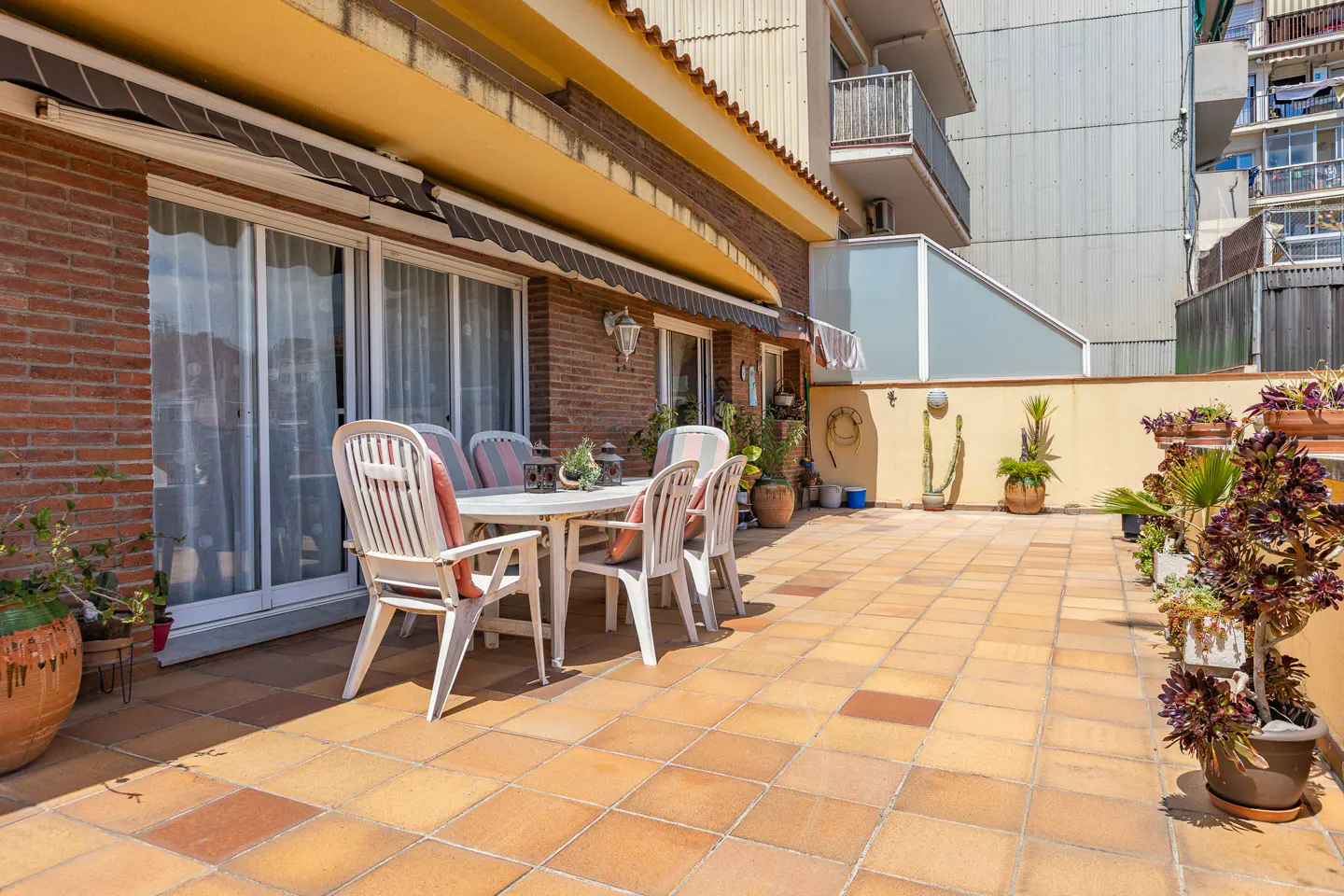 Outdoor patio with terracotta tiles, white table and chairs, brick wall, and potted plants.