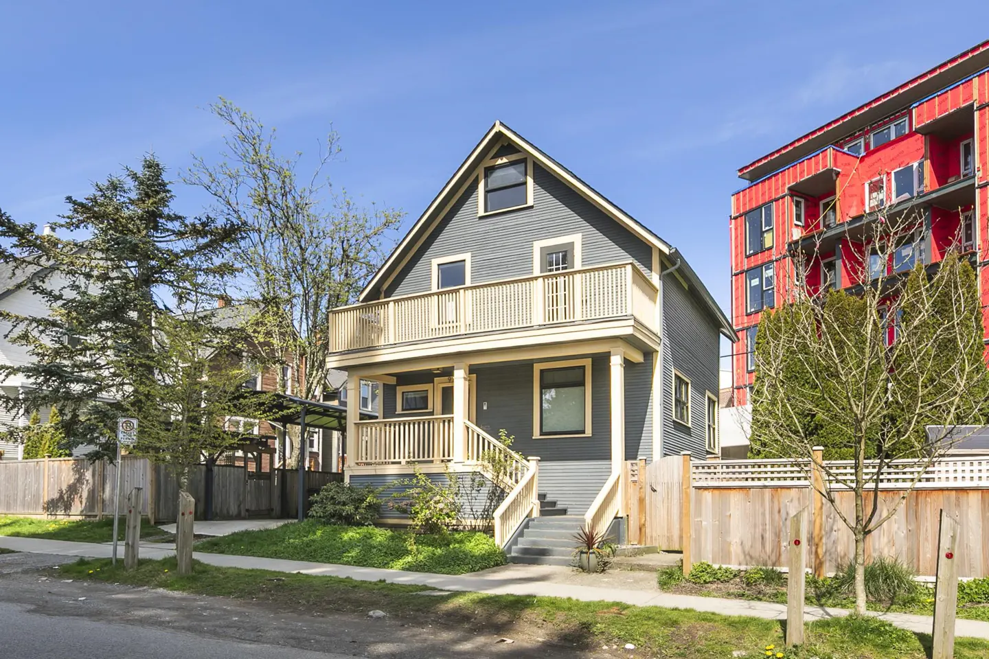 Two-story gray house with a cream-colored balcony and trim, surrounded by a wooden fence and greenery. A red building is visible in the background.