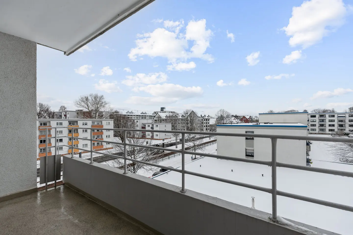 View from a balcony with a gray railing overlooking snow-covered buildings under a blue, cloudy sky.