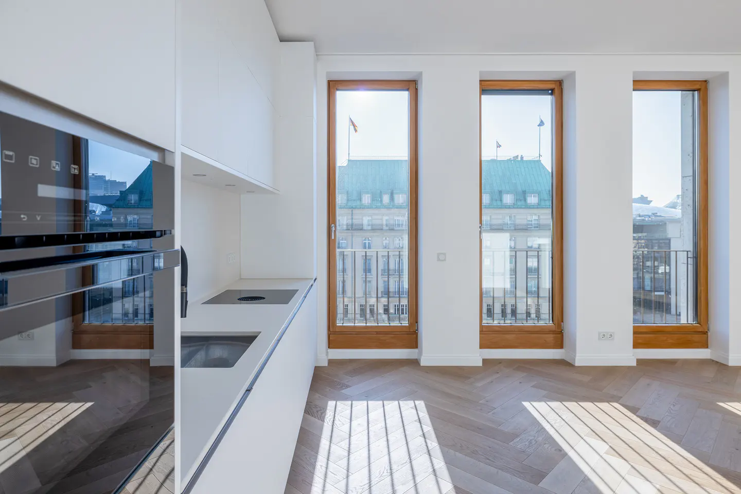 Bright, modern kitchen with white cabinets, black oven, and herringbone wood floors. Three windows offer a city view.