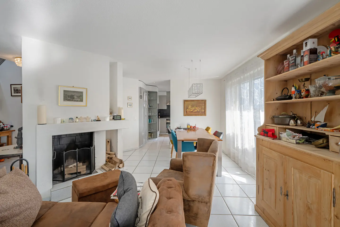 A bright living room with a brown sofa, fireplace, dining table, and a wooden cabinet filled with various items.