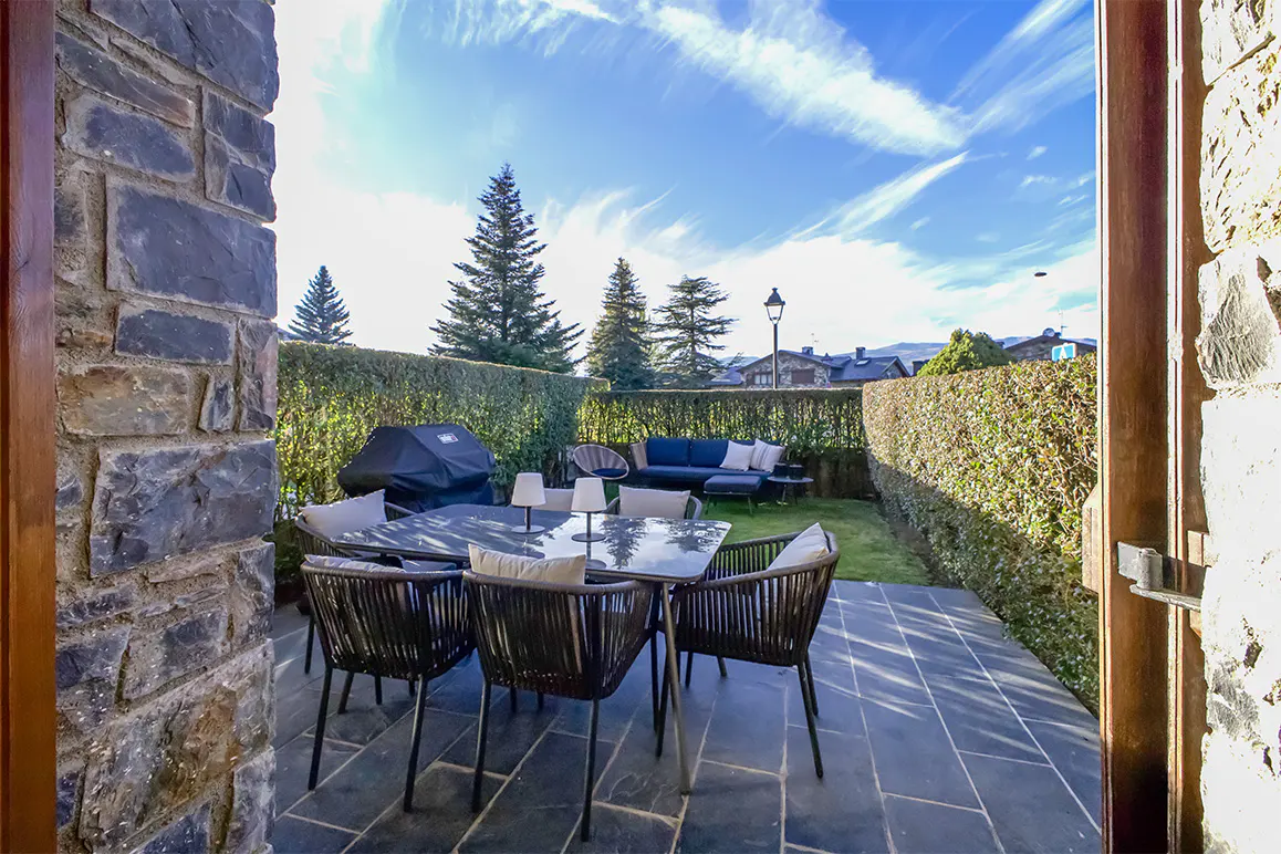 Outdoor patio with table, chairs, grill, and sofa, viewed from a stone doorway under a blue sky.